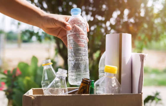 Mano sosteniendo una botella de agua junto a contenedores de reciclaje