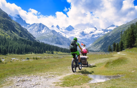 Familia en bicicleta y a pie por un prado alpino con nieve al fondo