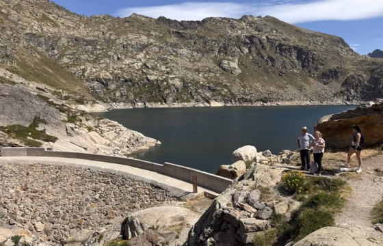 Lago de montana con el muro de una presa y tres personas en la orilla rocosa, paredes de granito al fondo
